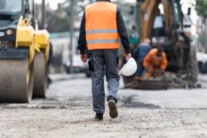 Construction worker walking through an active job site holding a hard hat near large machinery and roadwork.