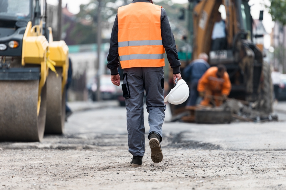 Construction worker walking through an active job site holding a hard hat near large machinery and roadwork.