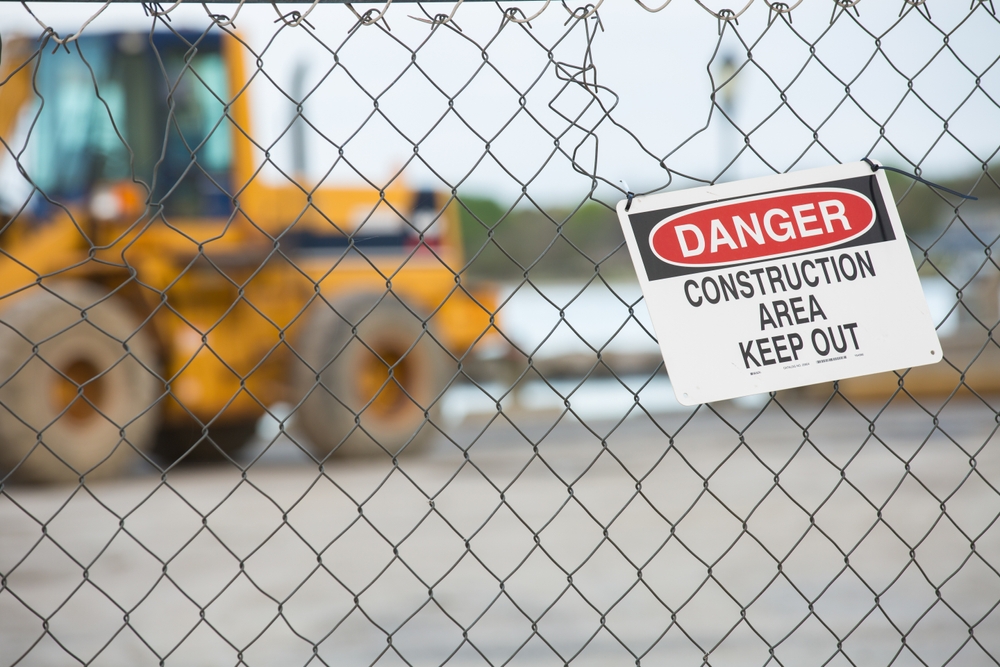 Danger sign reading “Construction Area Keep Out” posted on a chain-link fence with heavy equipment blurred in the background.