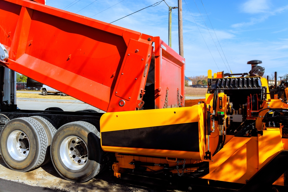 Dump truck unloading material beside heavy paving equipment at a construction site.